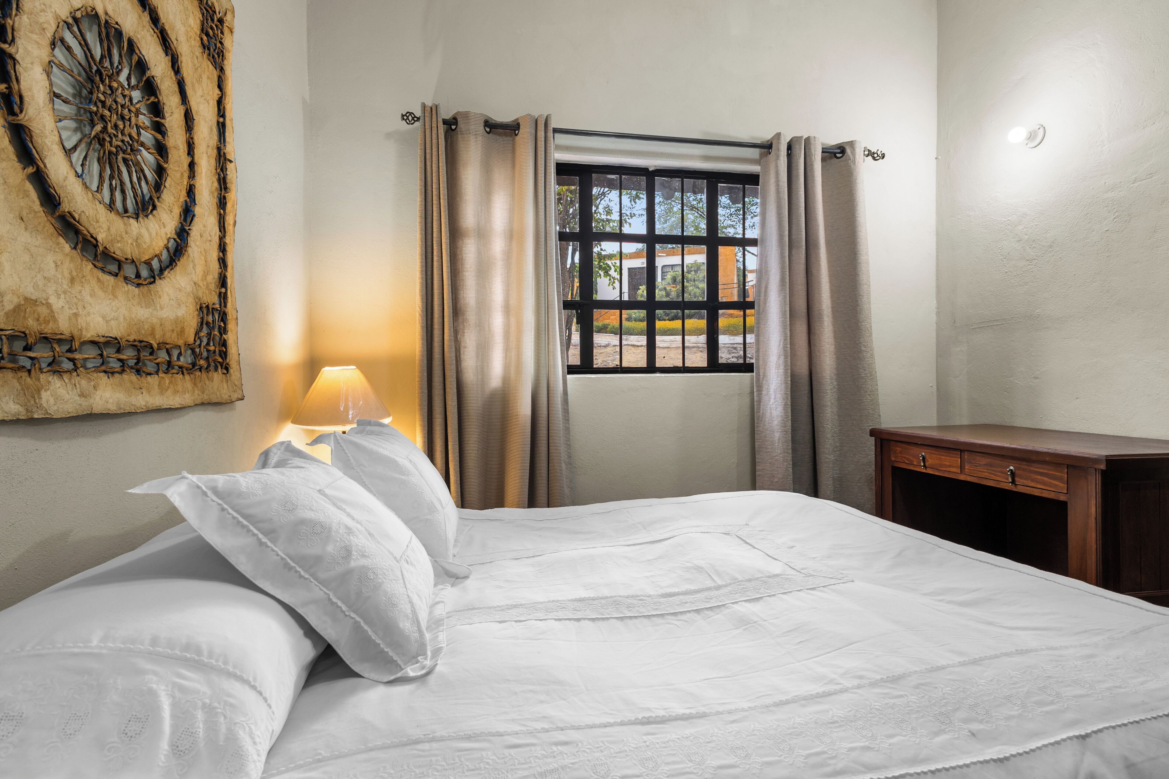 Una cama bien hecha con sábanas blancas, una lámpara de noche, una ventana con cortinas y una obra de arte en la pared.