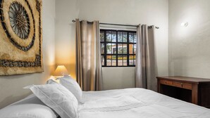 Una cama bien hecha con sábanas blancas, una lámpara de noche, una ventana con cortinas y una obra de arte en la pared.