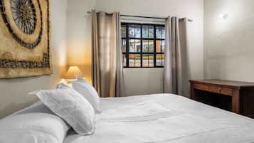 Una cama bien hecha con sábanas blancas, una lámpara de noche, una ventana con cortinas y una obra de arte en la pared.