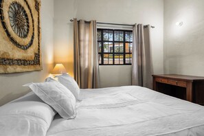 Una cama bien hecha con sábanas blancas, una lámpara de noche, una ventana con cortinas y una obra de arte en la pared.