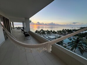 Un balcón con hamaca, sillas y vistas al atardecer sobre el mar.