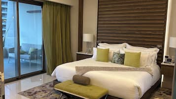 Habitación de hotel con cama, ventilador de techo, ventana grande con cortinas, una pequeña mesa con lámpara y vista a un balcón.