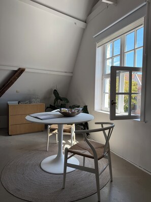 A room with a round table, chairs, a wooden cabinet, and a large window with a view of buildings. - Your home in Bruges, set in a historic setting just 5 minutes from the Main Market Square. (Brugge)