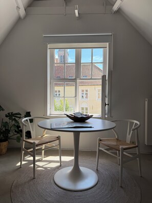 Ett rum med ett runt bord, två stolar, ett fönster med utsikt över byggnader och en växt.