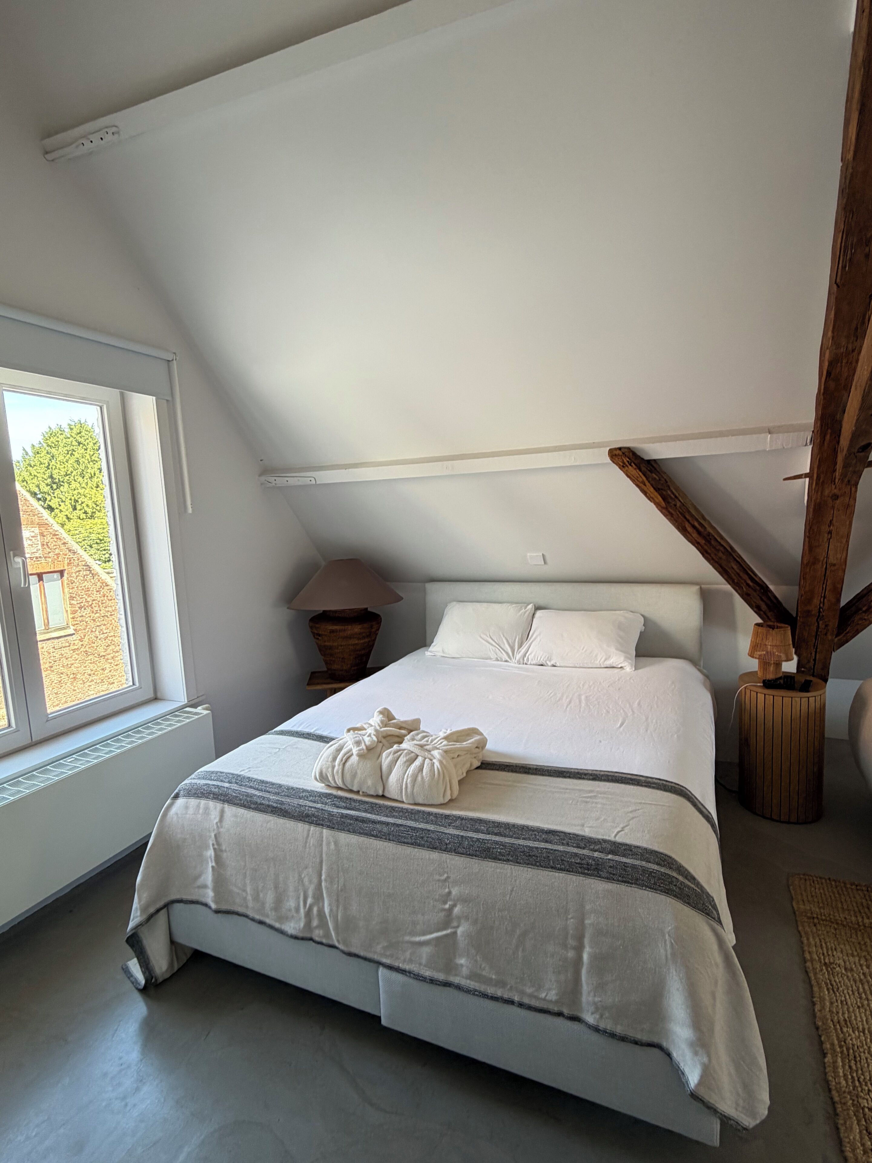 Un dormitorio con techo inclinado, cama con ropa de cama blanca, ventana con vista a un edificio y una lámpara sobre una mesita de noche de madera.