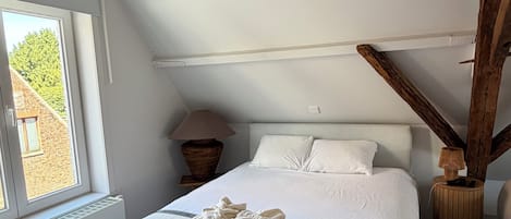 Un dormitorio con techo inclinado, cama con ropa de cama blanca, ventana con vista a un edificio y una lámpara sobre una mesita de noche de madera.