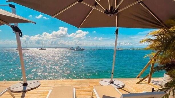 Eine Holzterrasse mit weißen Liegestühlen und einem großen Sonnenschirm mit Blick auf ein ruhiges Meer und Boote.