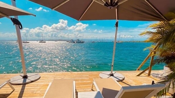 Una terraza de madera con sillones de descanso y sombrillas con vistas a un mar azul y despejado con barcos.