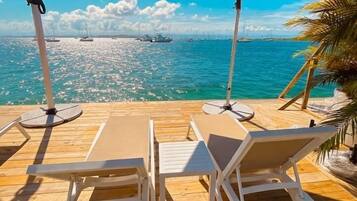 A wooden deck with lounge chairs and umbrellas overlooking a clear blue sea with boats.