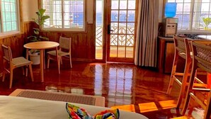 Habitación con suelo de madera, una mesa de comedor con sillas, una pequeña cocina y ventanas amplias con vistas al mar.