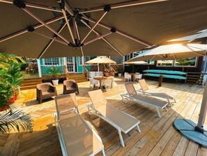 Une terrasse en bois avec des chaises longues et des parasols, entourée de plantes en pot et d’un espace piscine.