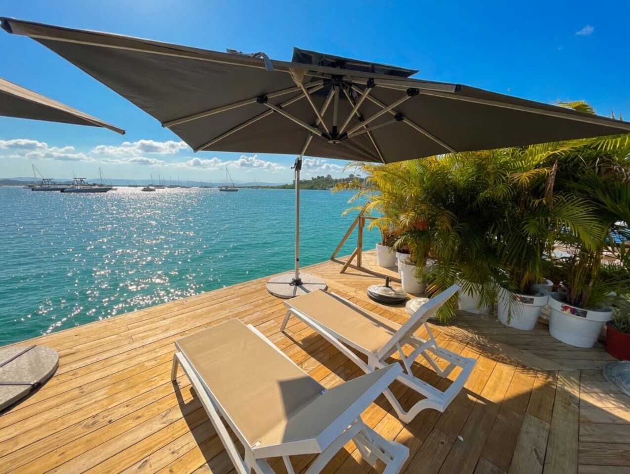 A wooden deck with lounge chairs and a large umbrella, overlooking a calm sea with boats in the distance.