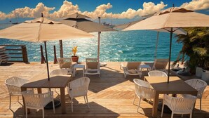 Eine Holzterrasse mit Tischen, Stühlen und Sonnenschirmen, mit Blick auf das Meer.