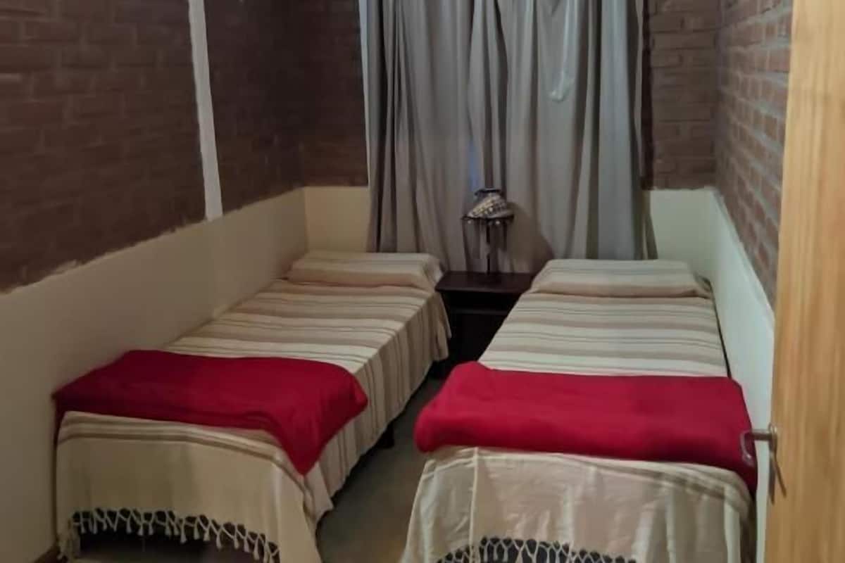 Una habitación con dos camas, un ventilador de techo, una ventana con cortinas y una lámpara sobre una mesa.