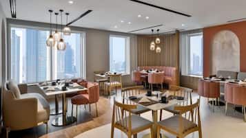 Un espace de restauration moderne doté de nombreuses tables et chaises, de grandes fenêtres offrant une vue sur la ville et d’un éclairage contemporain.