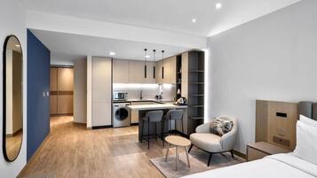 Quarto de hotel moderno com cama, escrivaninha e cadeira, pequena cozinha e banheiro.