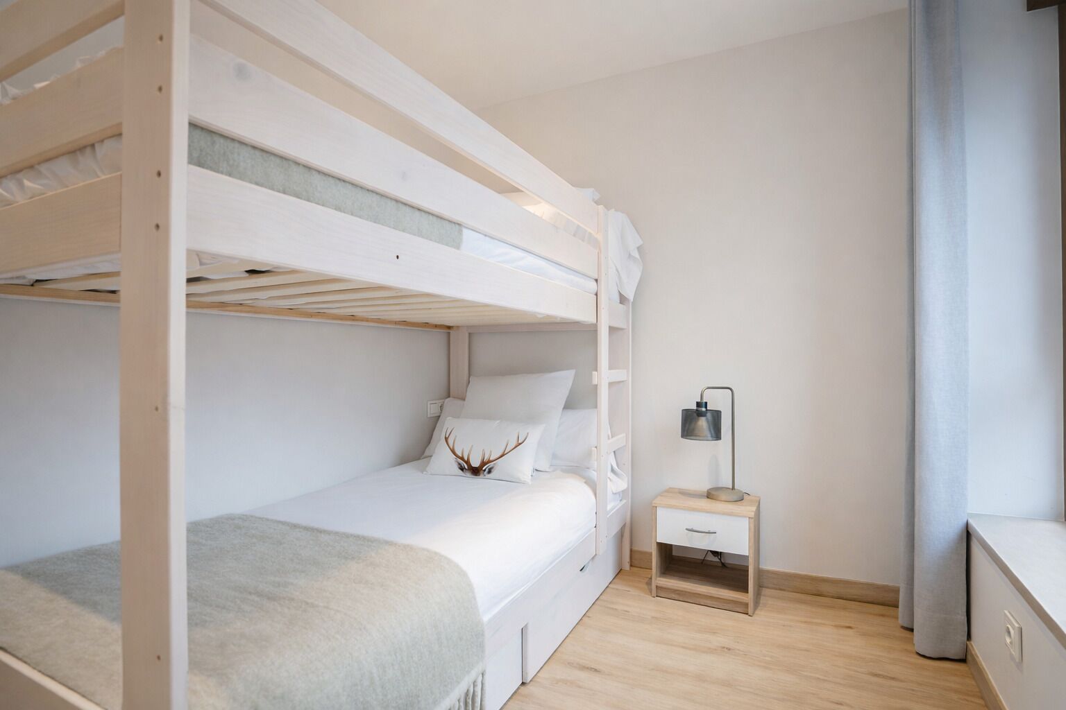 Une chambre avec un lit superposé, une table de chevet et une lampe, un plancher en bois et un mur uni.