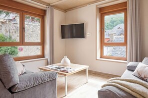 Ein Zimmer mit an der Wand montiertem Fernseher, einem Tisch mit Büchern, einer Couch und großen Fenstern mit Vorhängen.