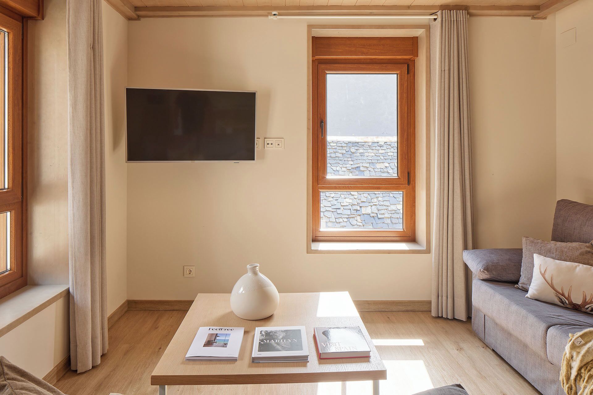 A room with a large window, a flat-screen TV, a table with books, and a sofa.