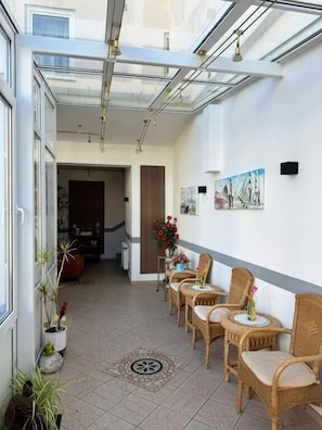 A sunlit corridor with wicker chairs, a glass ceiling, and a dining area with a table and chairs.