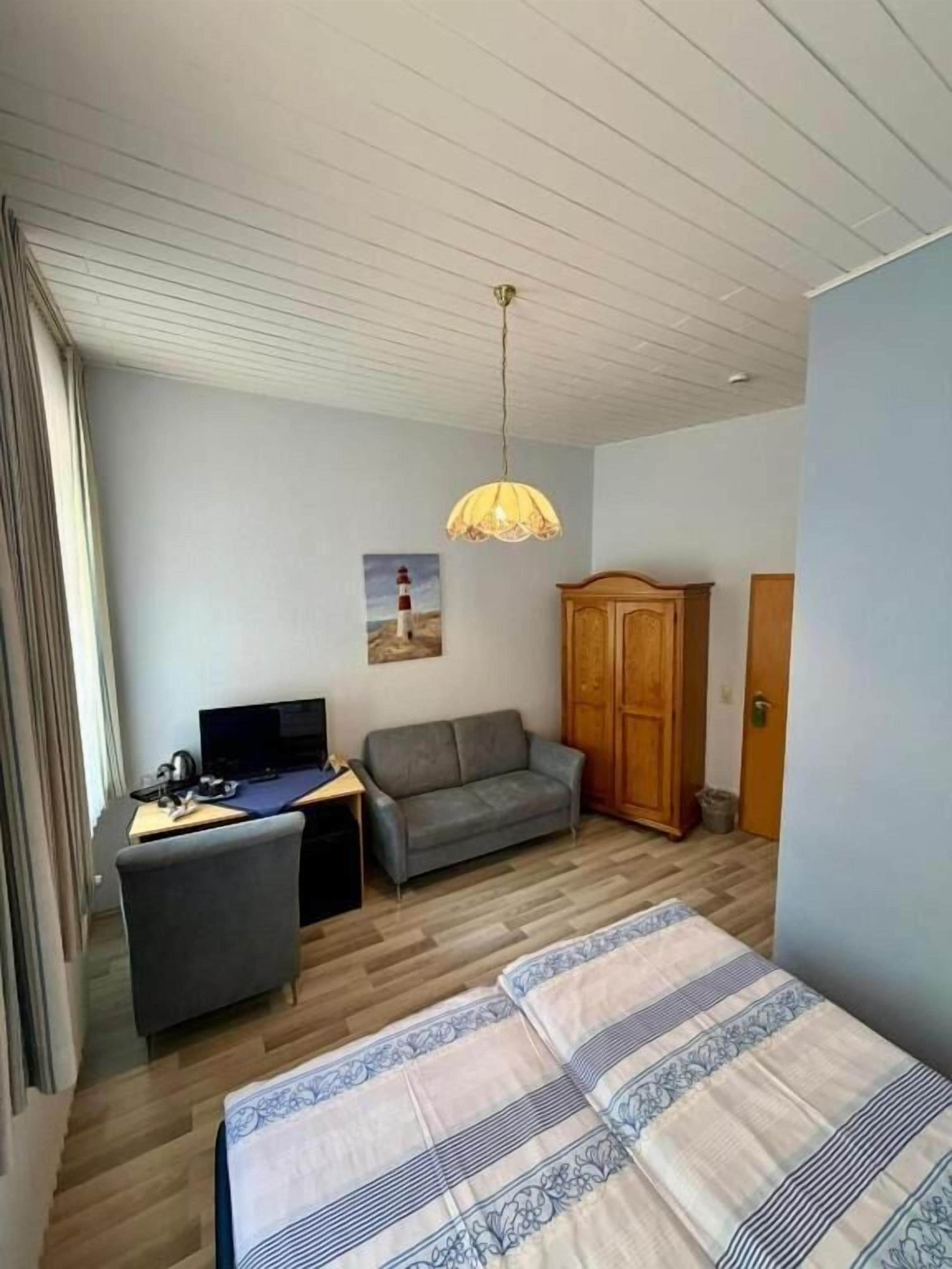 Ein Zimmer mit einem Bett, einem Schreibtisch mit Computer, einer Couch, einem Holzschrank und einer Pendelleuchte.
