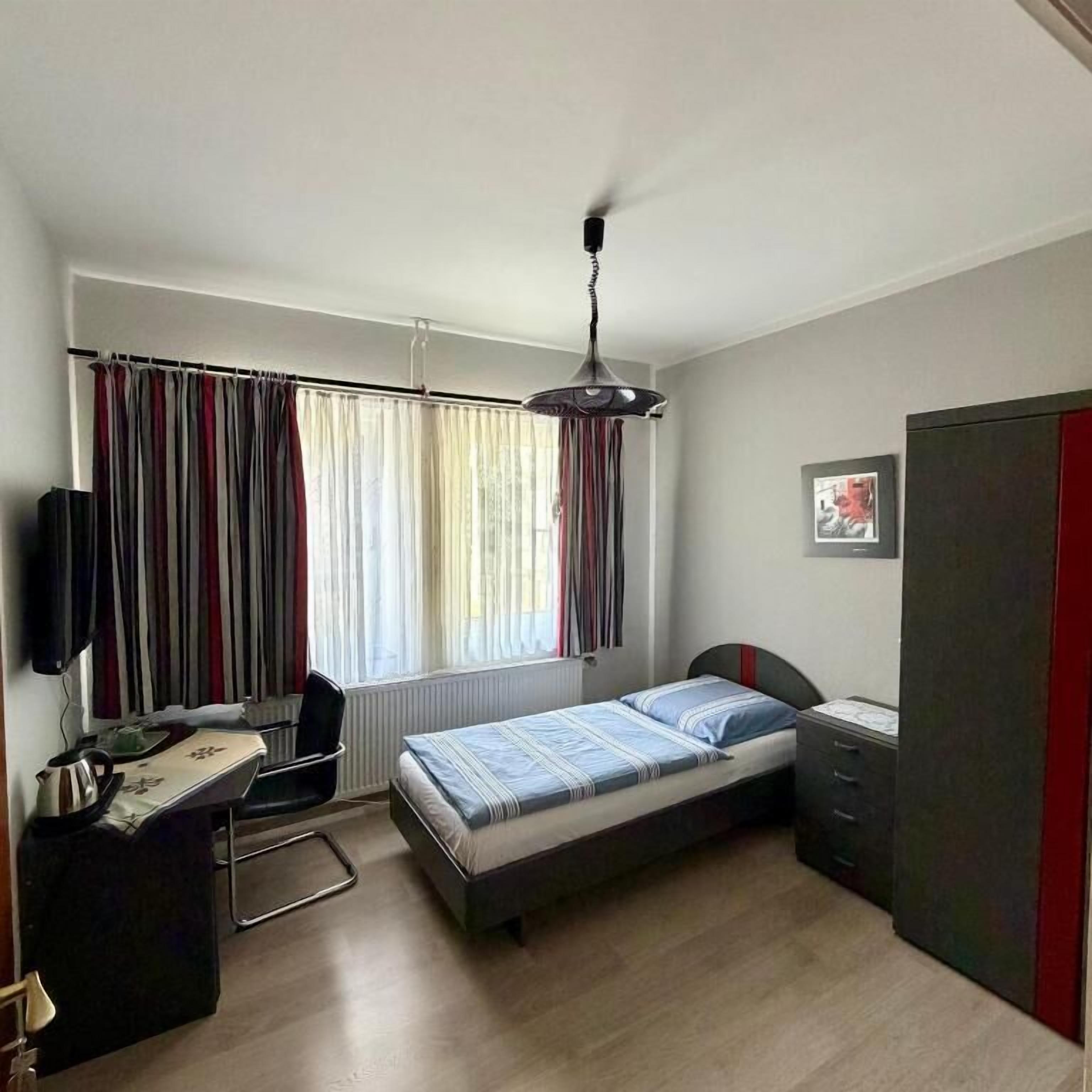 Quarto com uma cama, escrivaninha, cadeira, radiador, televisão e guarda-roupa.
