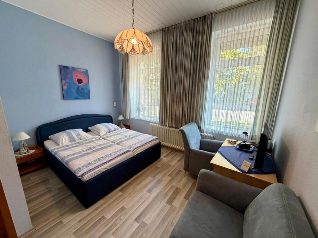 Un dormitorio con cama, escritorio, silla, lámpara, ventana con cortinas y un cuadro en la pared.