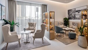 Ein modernes Wohnzimmer mit einem runden Tisch, zwei Sesseln, einer Couch, einem Bücherregal und einem großen Fenster mit Vorhängen.
