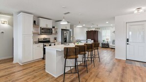 A modern kitchen with white cabinetry, a central island with bar stools, and a dining area visible in the background. - Al's Hideaway - Private Pool (Brunswick)