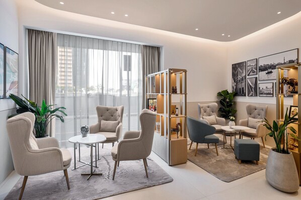 A modern living room with a round table, two armchairs, a sofa, a bookshelf, and a large window with curtains.