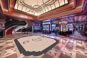A hotel lobby with a grand staircase, a bar area with slot machines, and a terrazzo floor with a name plaque.