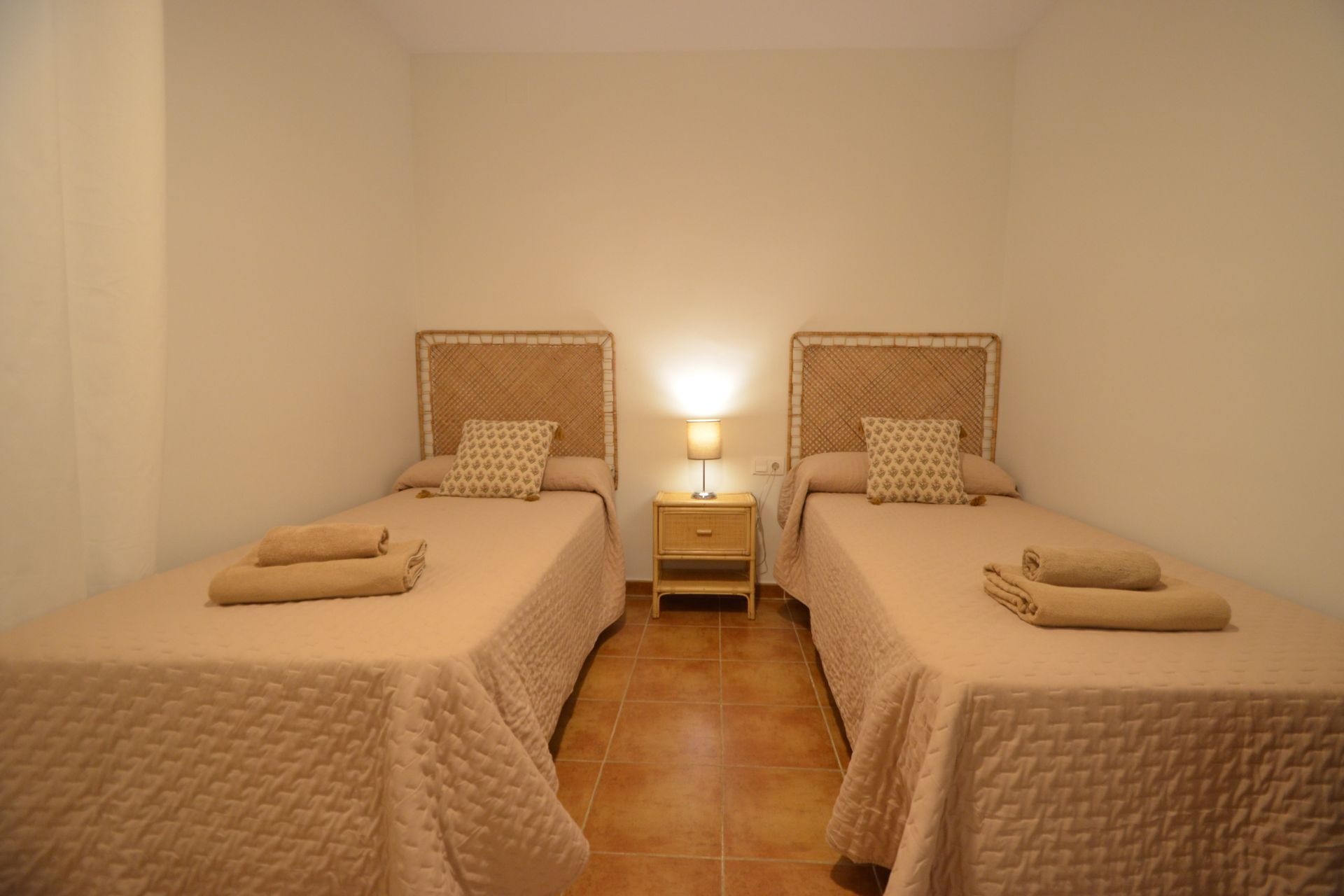 A room with two beds, each with a headboard, a nightstand with a lamp, and a tiled floor.