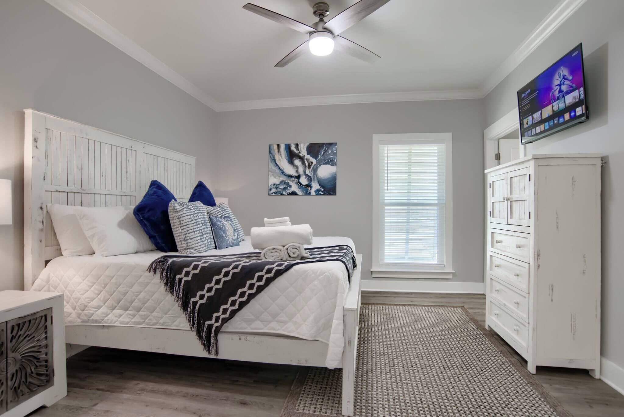 A bedroom with a bed, a ceiling fan, a TV, a dresser, and a window.