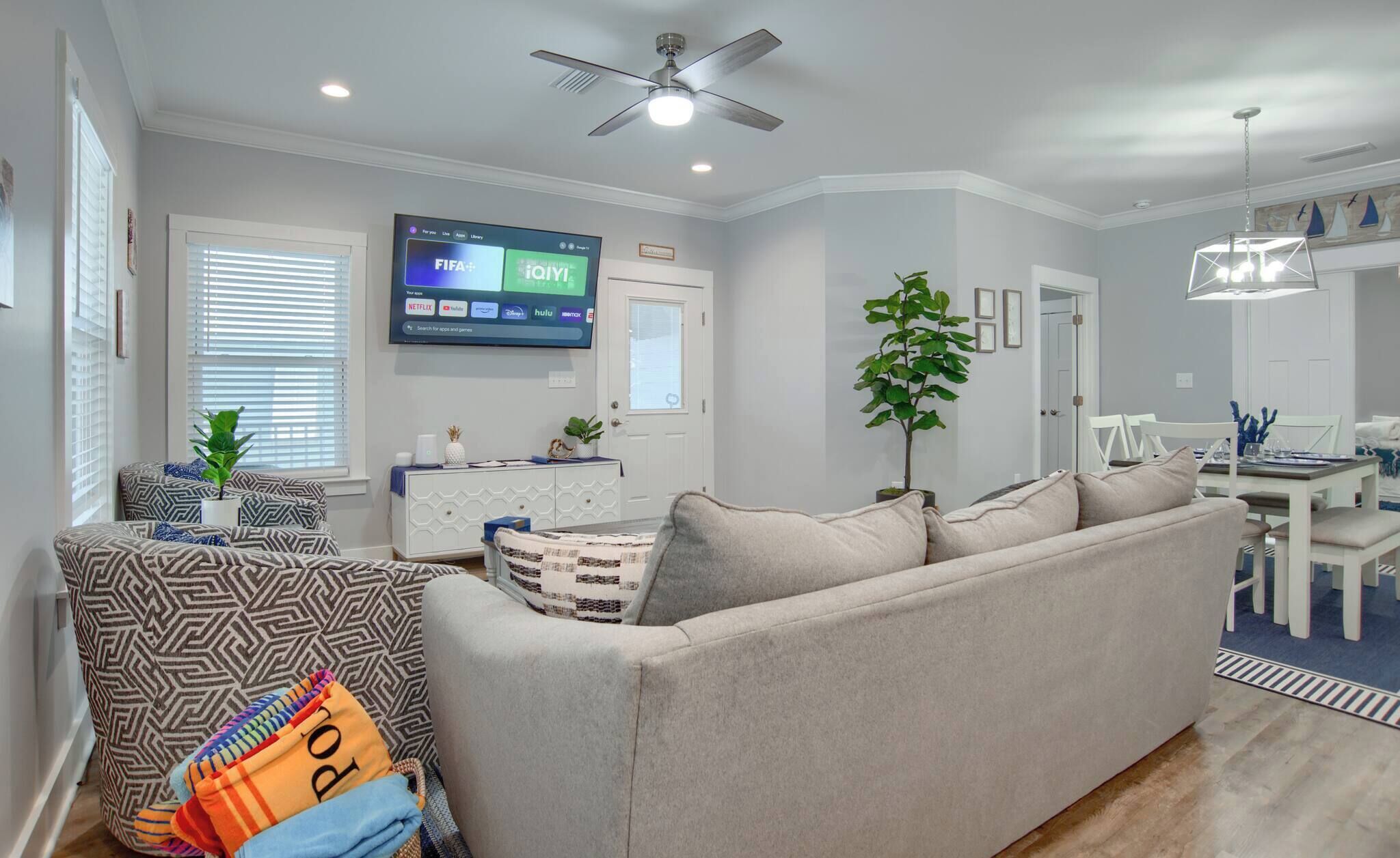 A living room with a sofa, a TV mounted on the wall, a ceiling fan, and a dining area with a table and chairs.