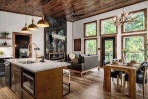 Eine moderne Küche mit einer großen Kücheninsel, ein Essbereich mit Holztisch und -stühlen, eine gemütliche Sitzecke mit einer Couch und große Fenster mit Blick ins Freie.