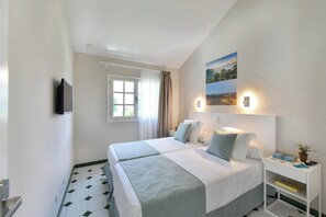 Ein Hotelzimmer mit zwei Betten, einem Fenster mit Vorhängen, einem Wandfernseher, einem Nachttisch mit Lampe und einem Wandbild.