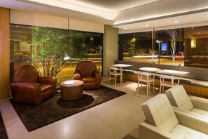 Uma área de estar moderna com poltronas de couro, uma mesa de centro redonda e um sofá. Janelas amplas oferecem vista para a rua.