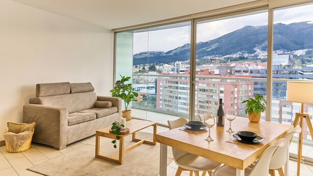 Una sala de estar moderna con sofá, mesa de comedor, sillas y un amplio ventanal con vista a la ciudad y a las montañas.