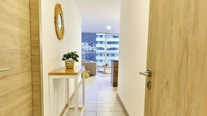 Un pasillo con una puerta de madera, una mesita con una planta y vistas a un balcón con un edificio al fondo.