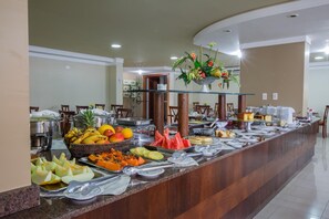 Buffet com diversas frutas e pratos, um expositor de vidro e uma área de estar com cadeiras.