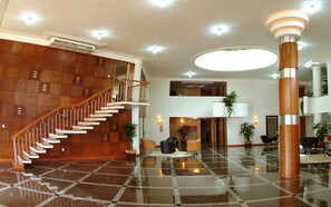 Um hall amplo com escadaria de madeira, uma coluna central imponente e piso xadrez.