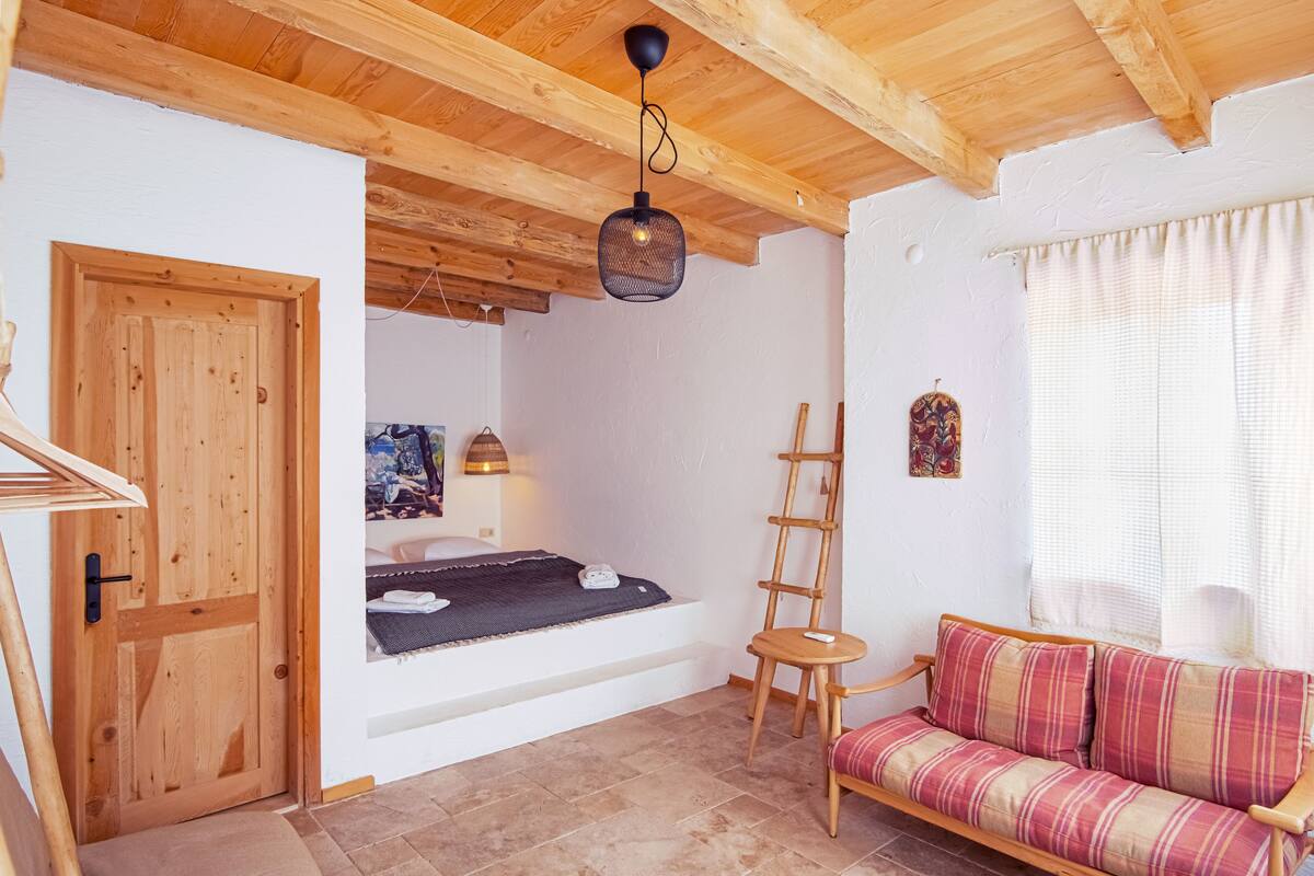 A cozy room with a bed, a wooden ceiling, a small table, a sofa, and a ladder.