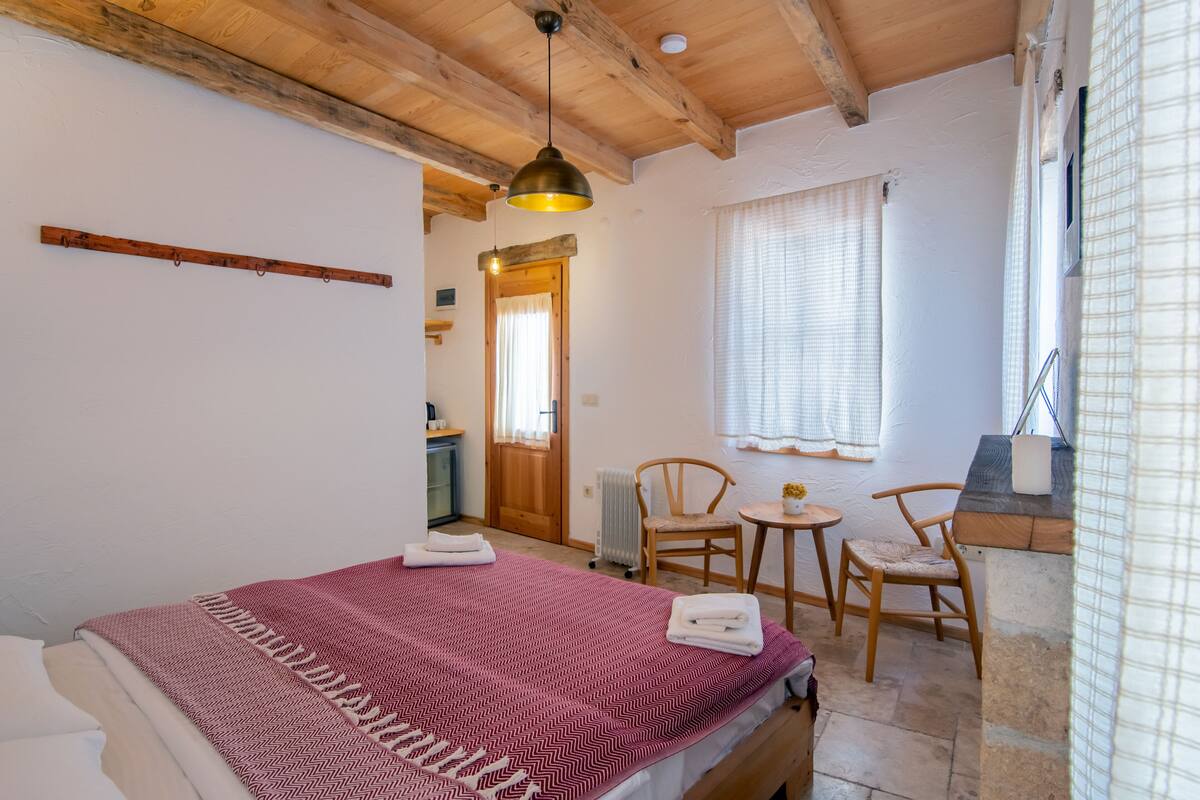 A bedroom with a bed, a wooden ceiling, a window with curtains, a small table, and a chair.