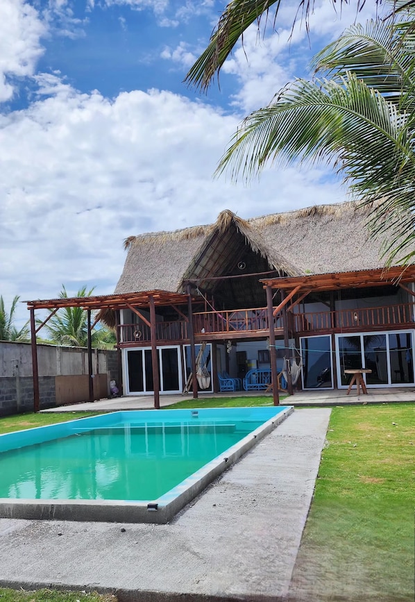 A pool with a wooden deck, a thatched-roof structure, and a view of palm trees.