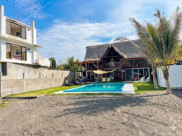 Una casa de dos pisos con piscina, una estructura de techo de paja y palmeras.