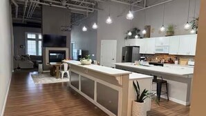 Una cucina moderna con mobili bianchi, un'isola centrale, illuminazione a sospensione e una TV in vista.