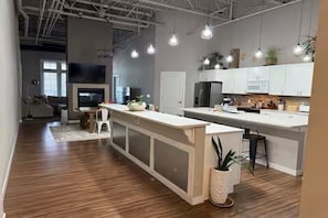 Une cuisine moderne avec des meubles blancs, un îlot central, des luminaires suspendus et un téléviseur bien en évidence.