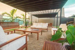 Un patio exterior con mobiliario de madera, una zona de estar techada y plantas en macetas.
