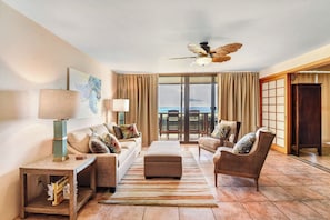 A hotel room with a sofa, armchair, ottoman, floor lamp, ceiling fan, and a view of the ocean through large windows. - NEW Kauai Oceanview - Spacious, AC, Pool, Sea Views (Koloa)