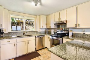 A kitchen with white cabinets, granite countertops, a dishwasher, an oven, and a window with a view of palm trees. - NEW Kauai Oceanview - Spacious, AC, Pool, Sea Views (Koloa)